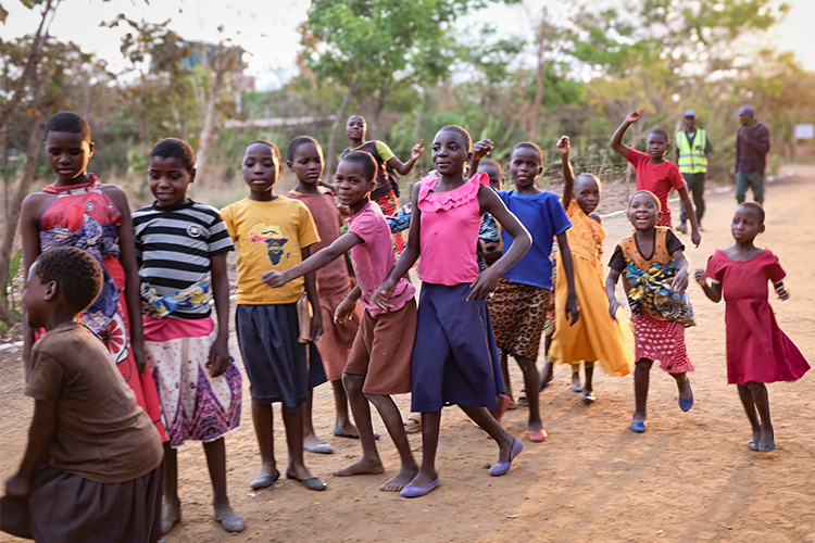 Malawian childern in a group outside with trees and a security guard in the background.