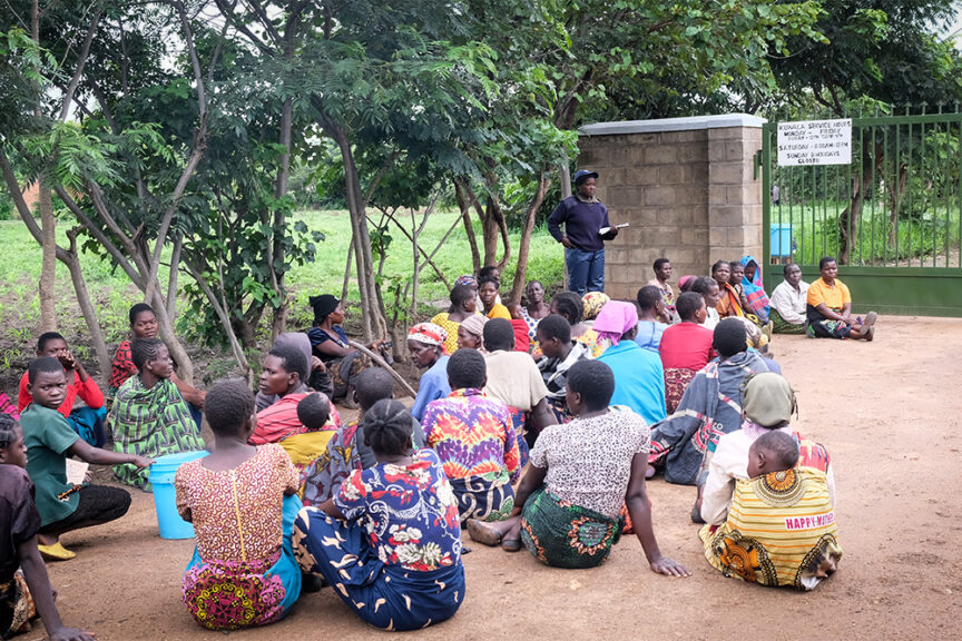 Group of villagers siting down at Kuwala's main gate with the security guards speaking to the group before they enter the Campus. There are trees in the background.