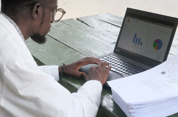 Teacher Martin is outside on a green table entering student papers on a laptop.